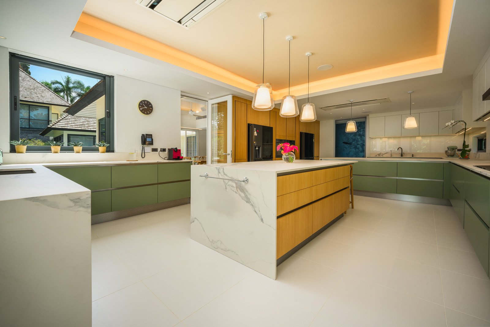 Modern kitchen with green cabinets, white marble island, and wooden accents, with pendant lights and a window showing a house and palm trees outside.