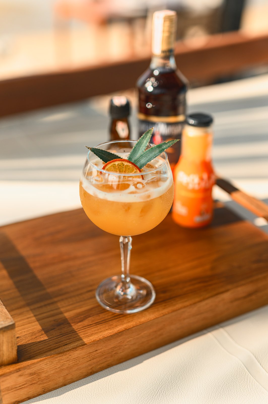 A cocktail glass with a yellow-orange drink garnished with a slice of orange and pineapple leaves, on a wooden serving board.