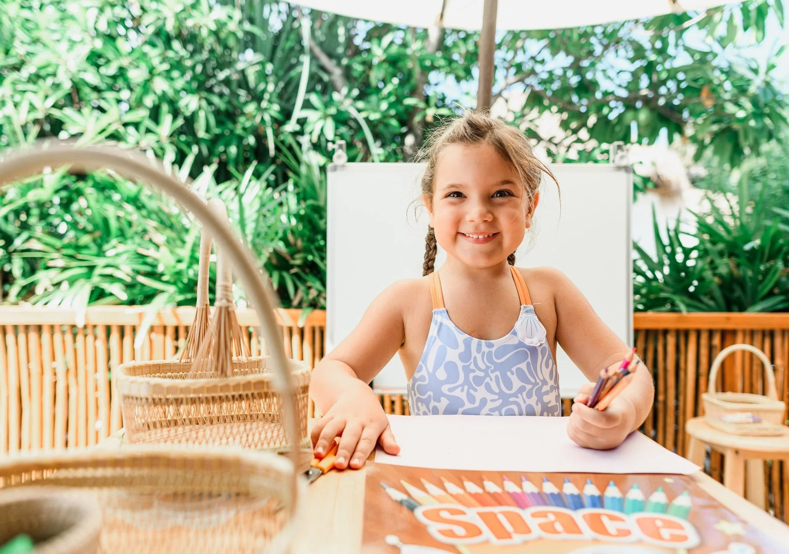 A young girl with braided hair, smiling, sitting at a table outdoors, surrounded by greenery, holding colored pencils, with art supplies and colored paper on the table.