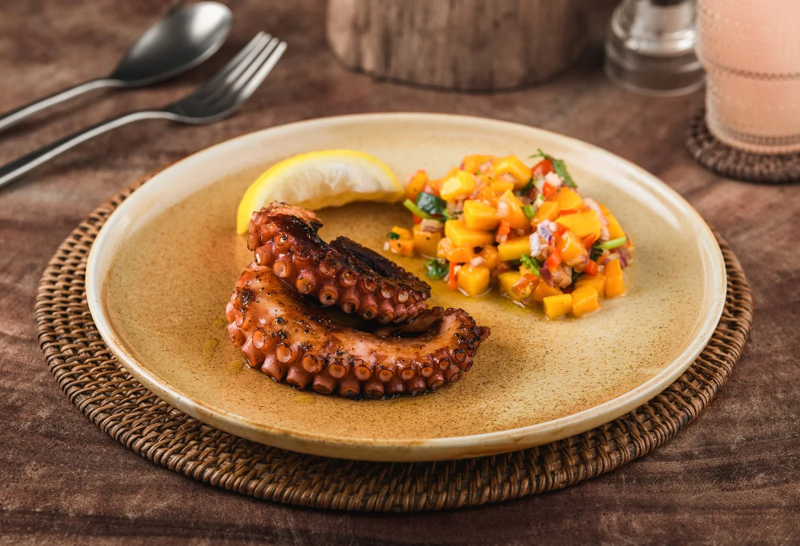 A plate with grilled octopus tentacle, lemon wedge, and a side of mango salad, on a woven placemat on a wooden table.