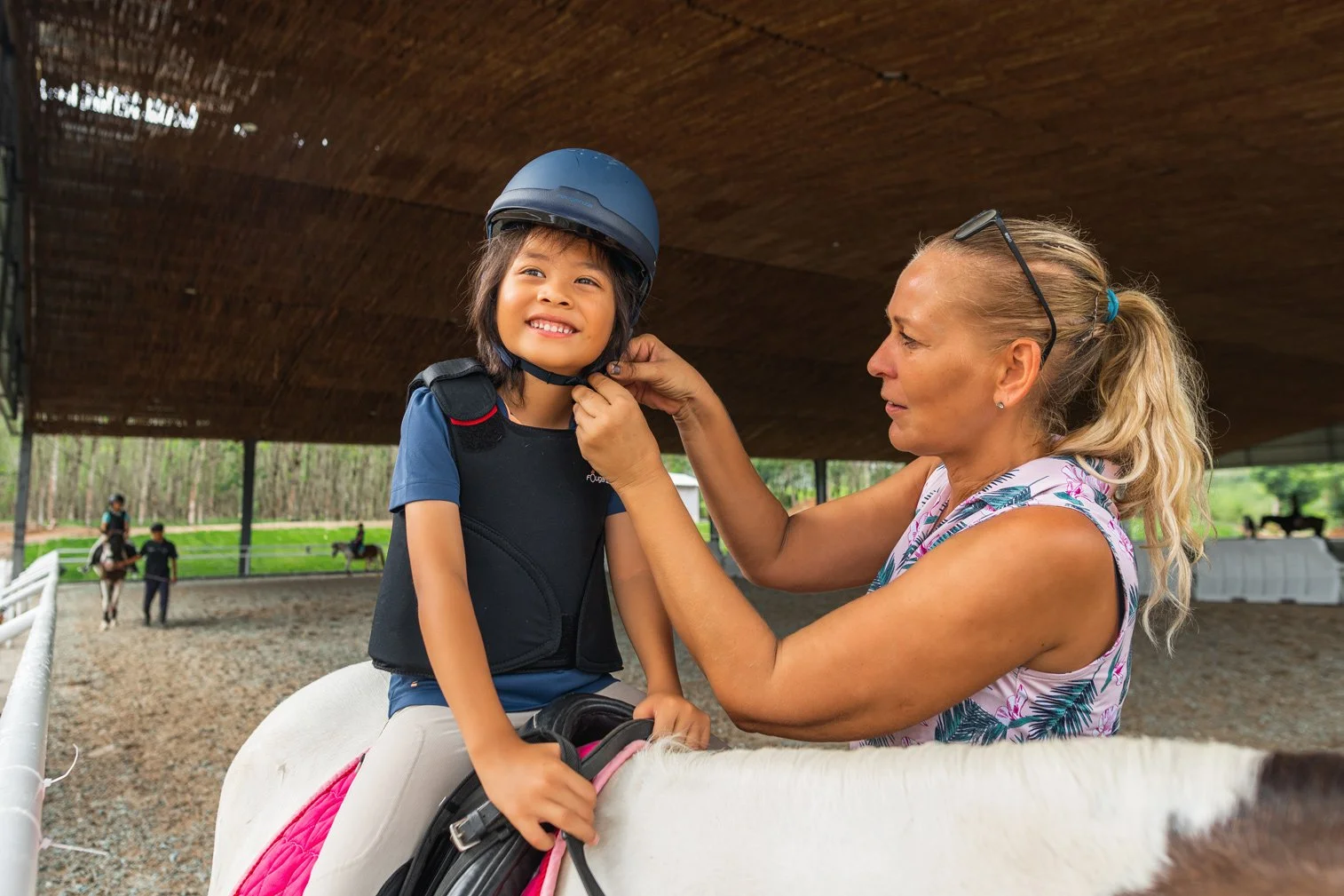 A girl is getting her helmet fitted by a woman on horseback at an outdoor riding arena.