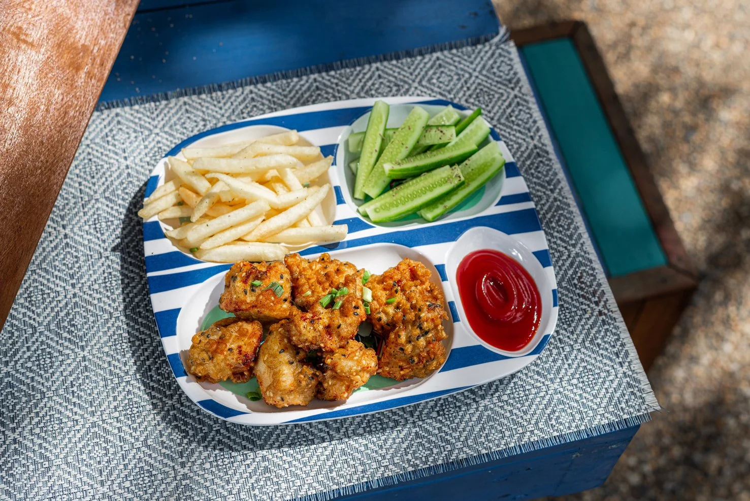 A divided tray with French fries, cucumber sticks, fried chicken pieces, and ketchup on a table outside.
