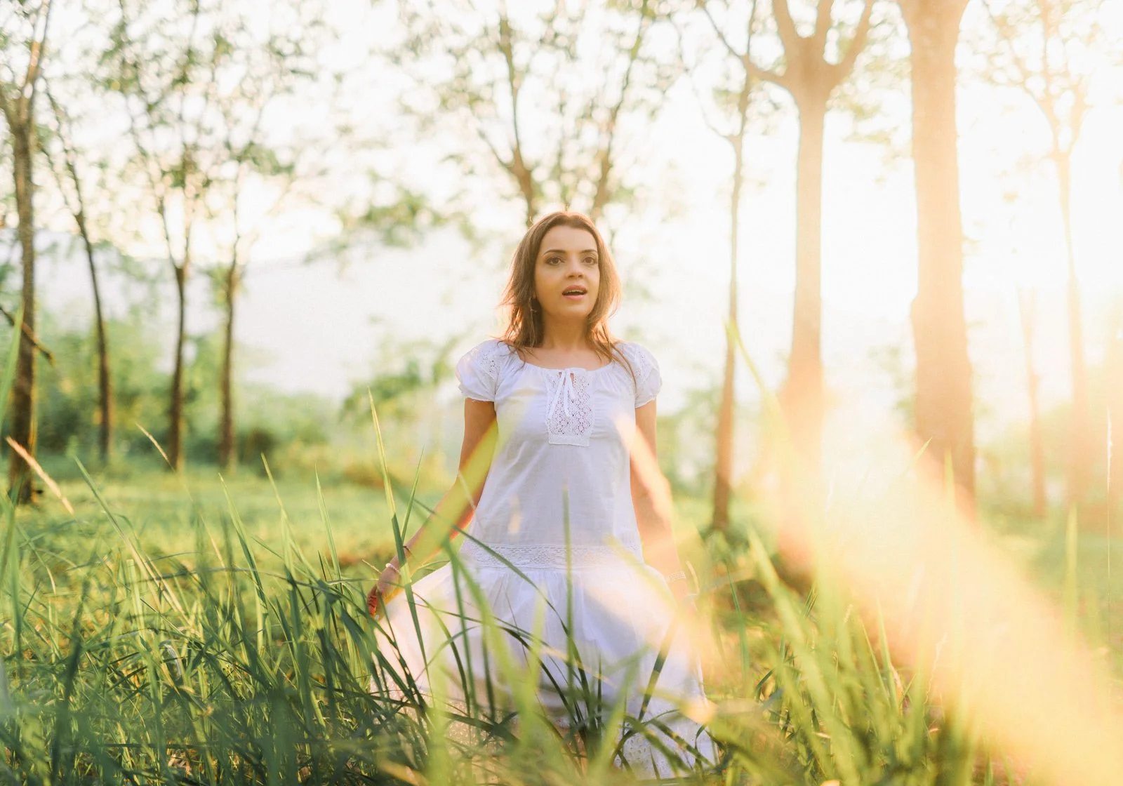 A woman wearing a white dress standing in a lush green field with tall grass, during a sunny day with bright sunlight filtering through trees in the background.