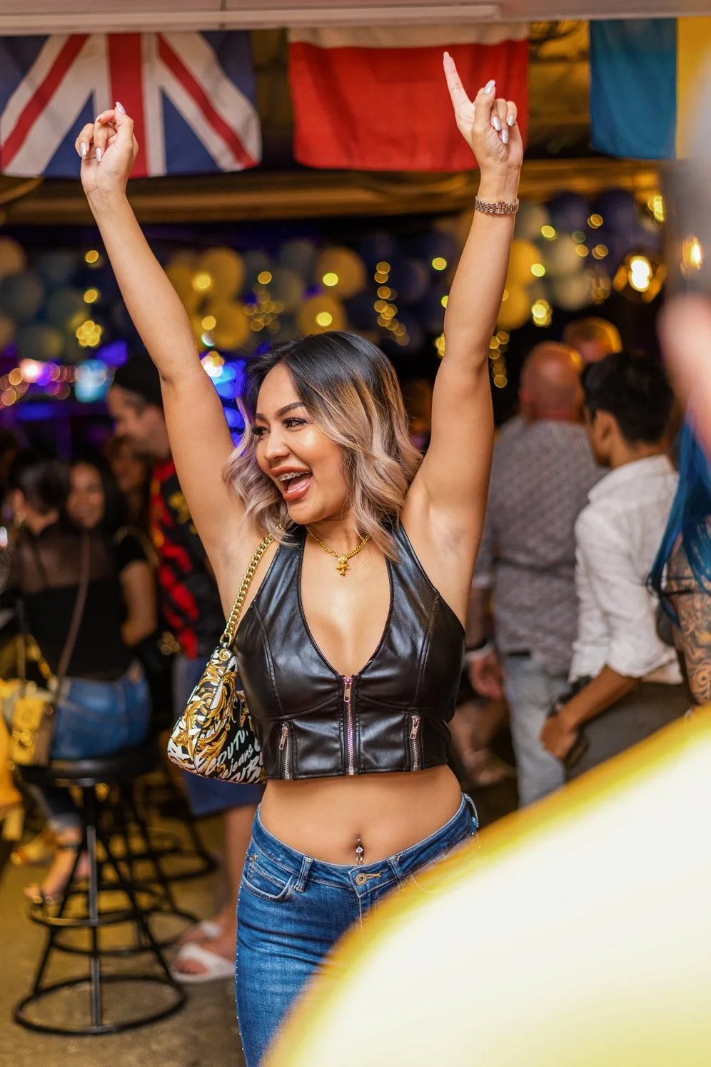 A young woman at a lively party or club, dancing with arms raised, smiling, with a background of colorful balloons and flags, including the Union Jack.