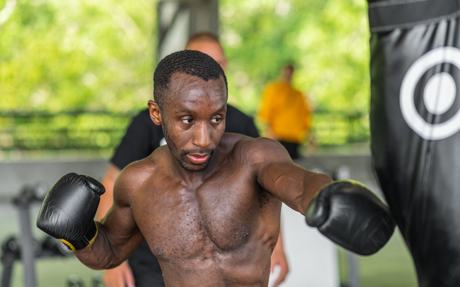A shirtless male boxer with black gloves practicing punches on a punching bag in a gym. He is sweating and focused, with a trainer or companion in the background. The gym has a green outdoor scene visible through windows.