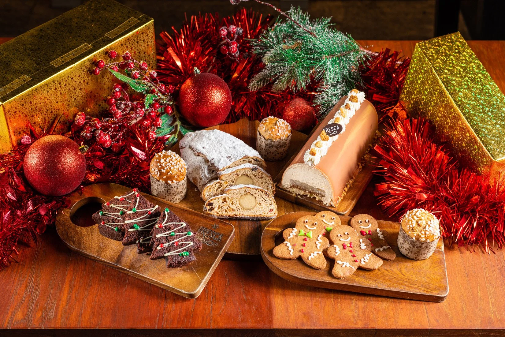 Decorative Christmas feast includes gingerbread cookies, a Yule log cake, a fruitcake, Christmas ornaments, tinsel, and holiday greenery arranged on wooden platters and a table, surrounded by gold and red gift boxes and festive decorations.