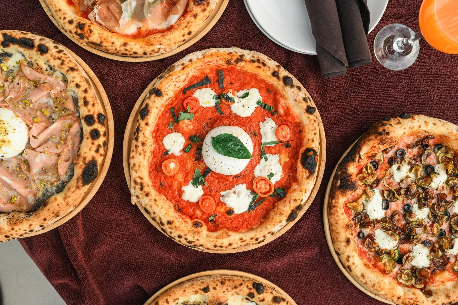 Assorted pizzas on a dark red tablecloth, including a Margherita with basil and mozzarella, and other pizzas with various toppings like ham, olives, and roasted vegetables.