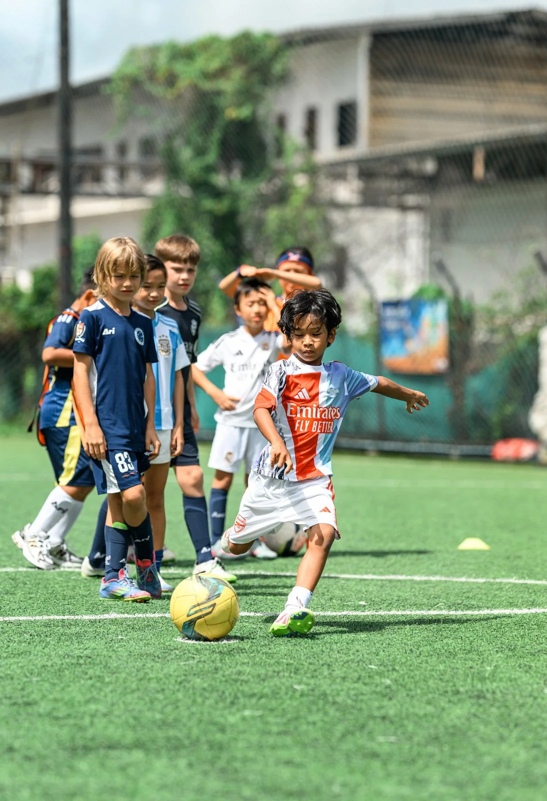 Young boy in a white and orange soccer uniform kicking a yellow soccer ball during a youth soccer practice on a green field with other children watching.