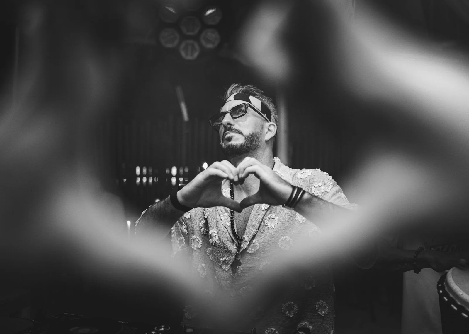 A man with dark hair, beard, and sunglasses making a heart shape with his hands. He is wearing a patterned shirt, a headband, and necklaces, and appears to be at a music or dance event with stage lighting in the background.