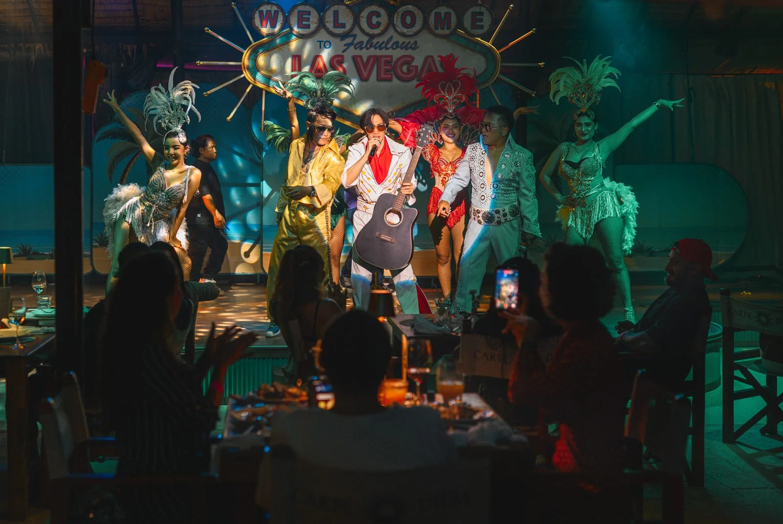 Performance on stage at a Las Vegas-style show with dancers in glamorous costumes and a lead performer with a guitar, audience seated at tables watching and enjoying the show.