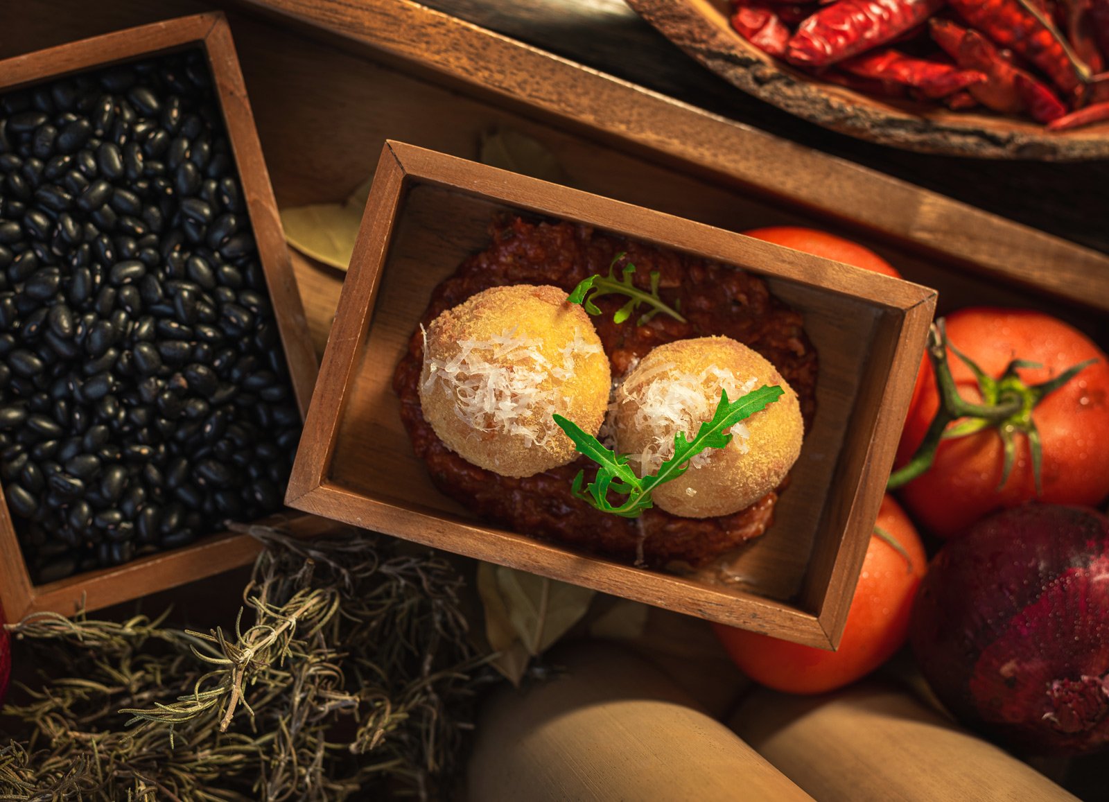 Three fried cheese spheres garnished with shredded cheese and parsley, inside a small wooden box, surrounded by black beans, tomatoes, and herbs.