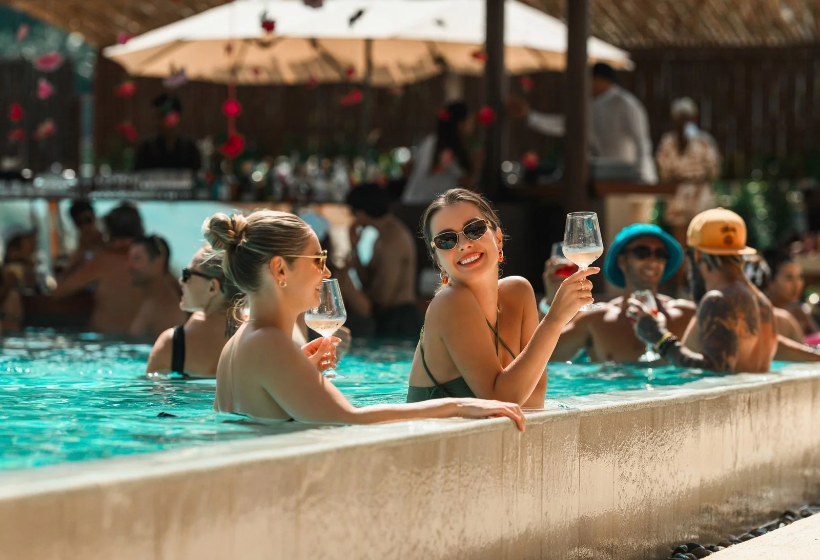 People enjoying drinks in a swimming pool at a lively outdoor party or bar, with people wearing sunglasses and colorful hats, surrounded by a wooden fence and umbrella in the background.