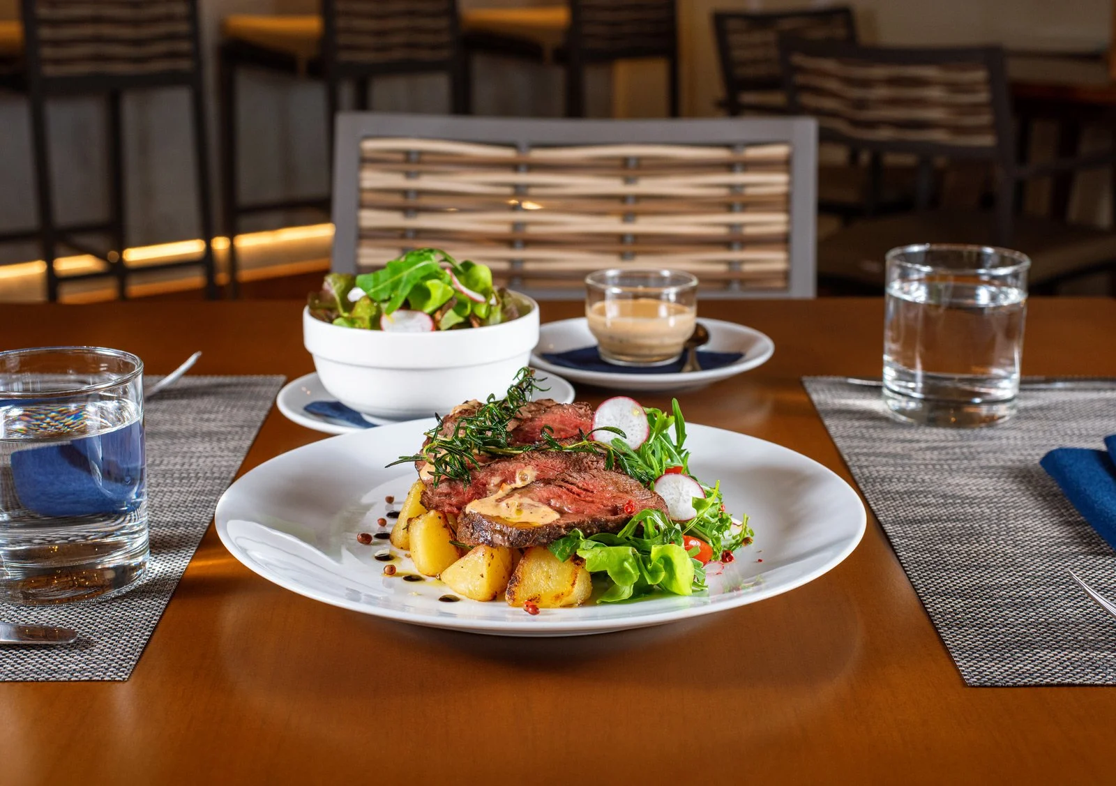 A plated meal with slices of steak, roasted potatoes, and fresh greens, accompanied by a side salad, on a wooden dining table with glasses of water, napkins, and small bowls of dressing or sauce in a restaurant setting.