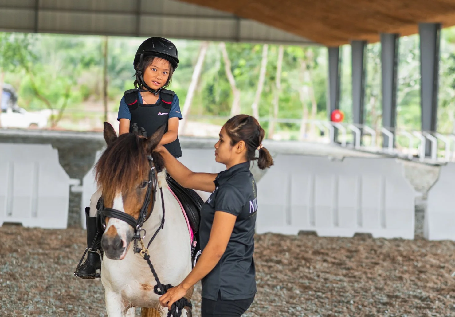 A girl wearing a helmet and safety vest is sitting on a horse in an indoor riding arena. A woman is standing beside the horse, holding its reins and adjusting the girl’s vest. The arena has a covered roof with large windows and trees visible outside.