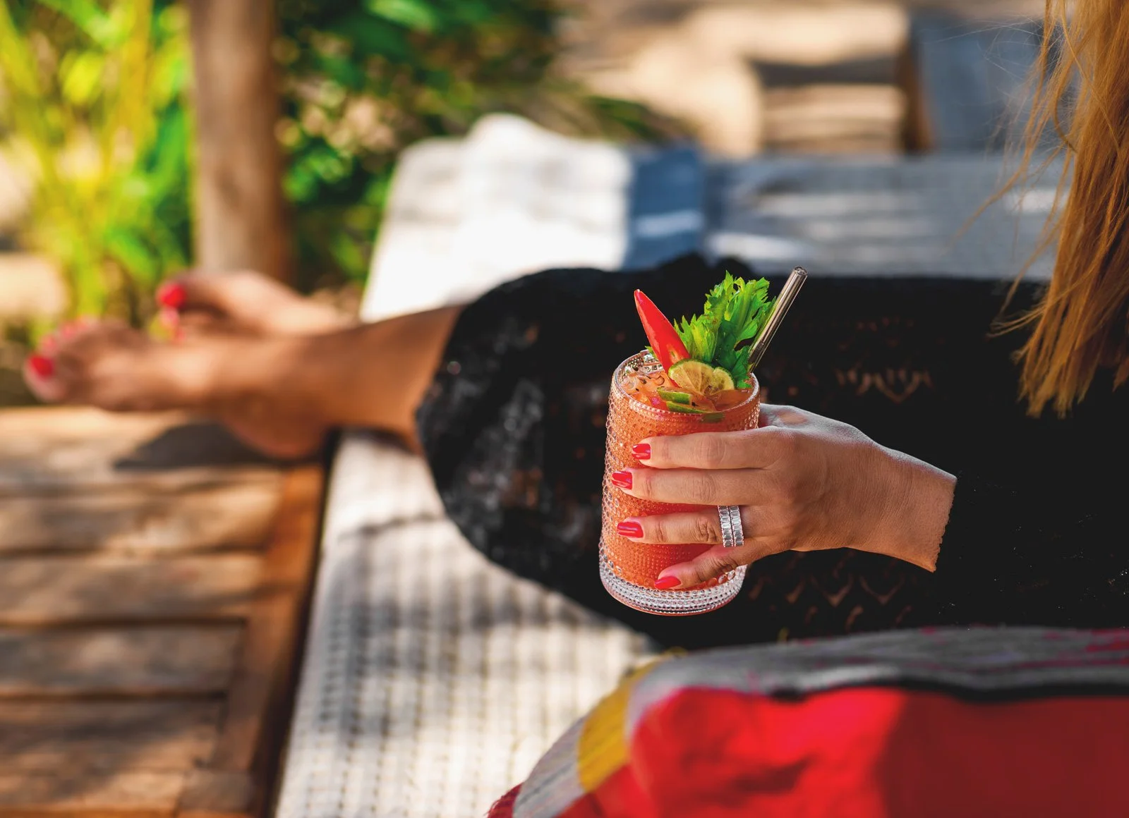 A person with red painted nails and a large ring on their finger is holding a colorful cocktail garnished with lime slices, a red chili pepper, and green herbs. The person is sitting outdoors on a wooden bench with greenery in the background.