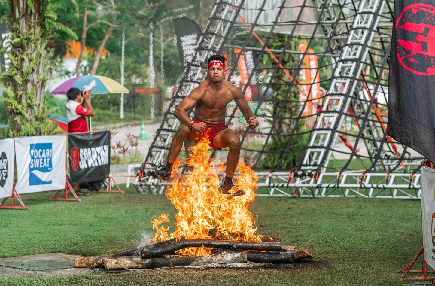 Man wearing a red headband and shorts jumping over fire on logs during an obstacle course event.