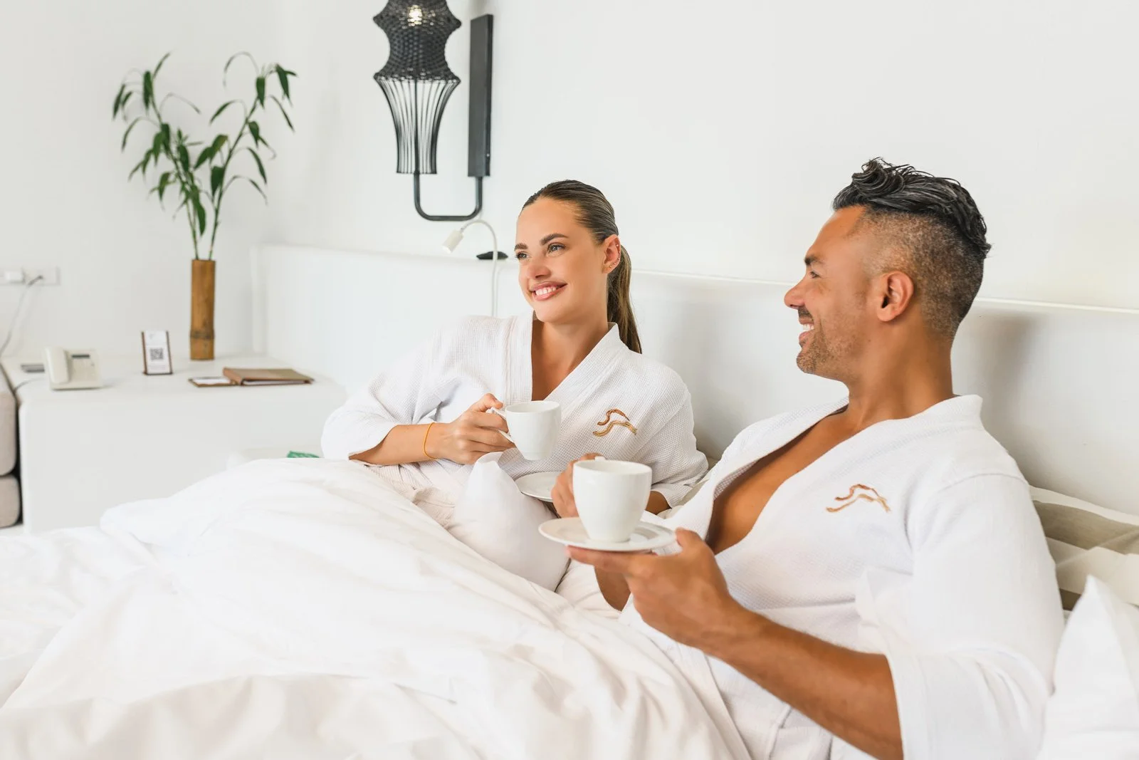 A man and woman sitting up in bed, wearing white robes, smiling and holding coffee cups, in a bright and modern bedroom with minimal decor.