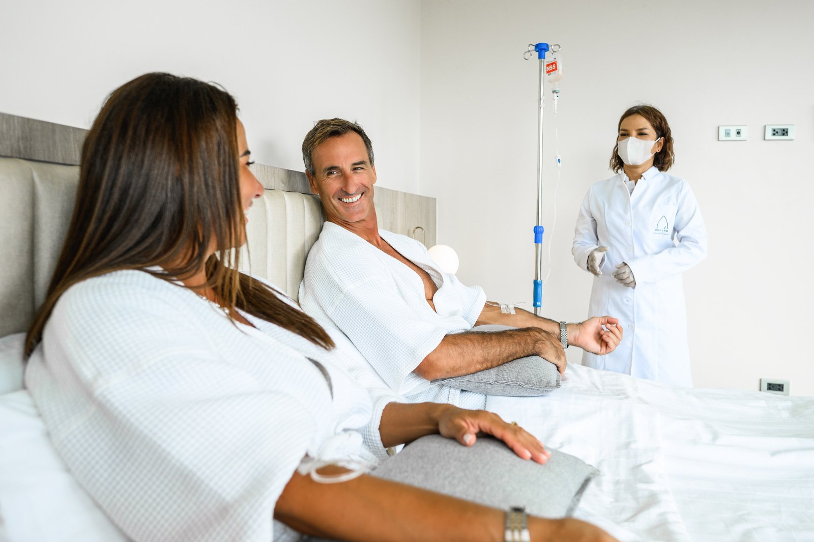 A group of three people in a hospital room. A woman is lying in bed, a man sitting next to her and smiling, and a nurse standing nearby in a white coat and face mask.