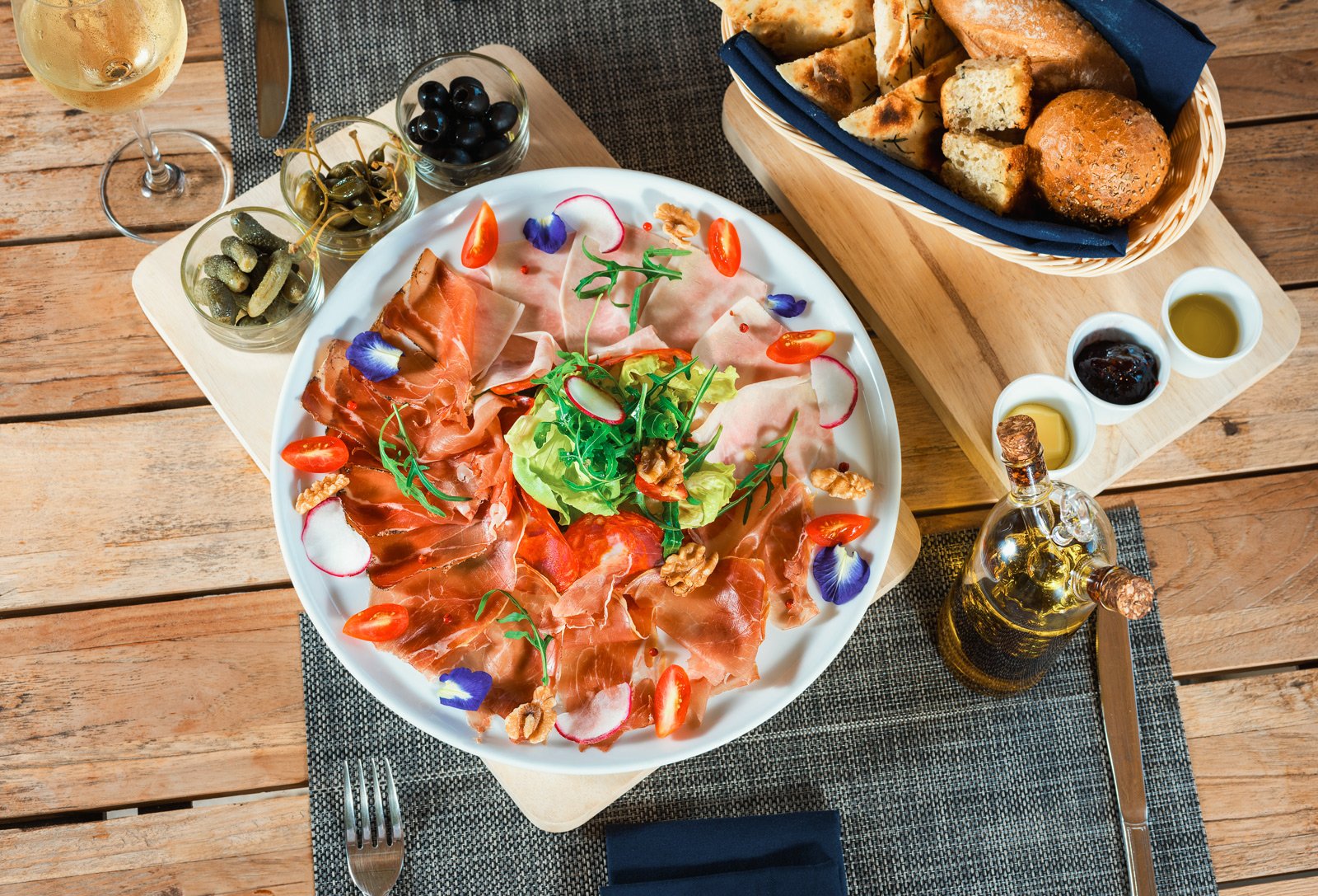 A round white plate of assorted cured meats, garnished with cherry tomatoes, radish slices, arugula, and edible flowers, served on a rustic wooden table with a glass of white wine, small bowls of pickles, olives, and condiments, alongside a basket of