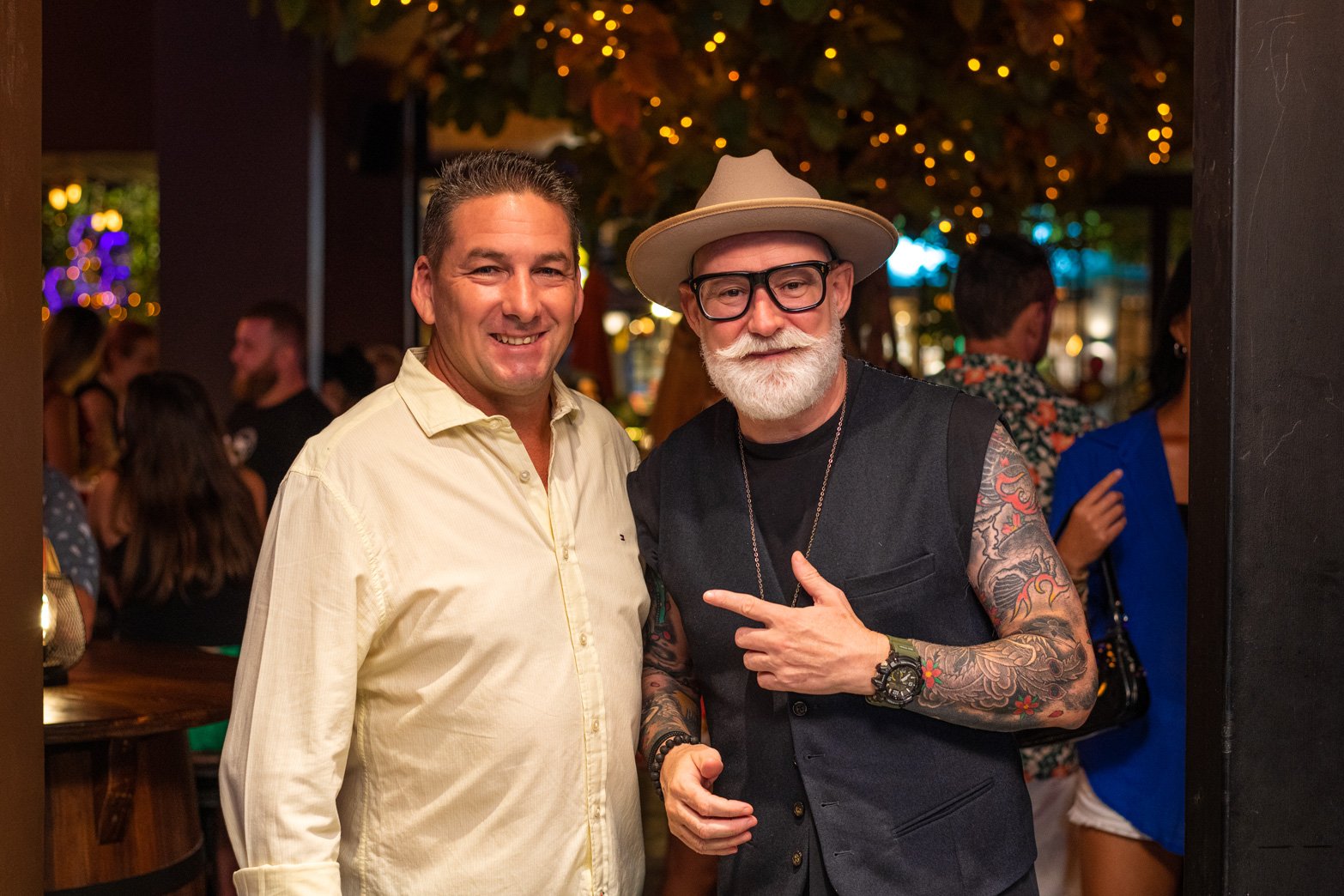Two men standing together at a lively indoor event, smiling at the camera. One man is wearing a light-colored shirt, and the other is dressed in black with a hat, glasses, and has tattoos on his arms.