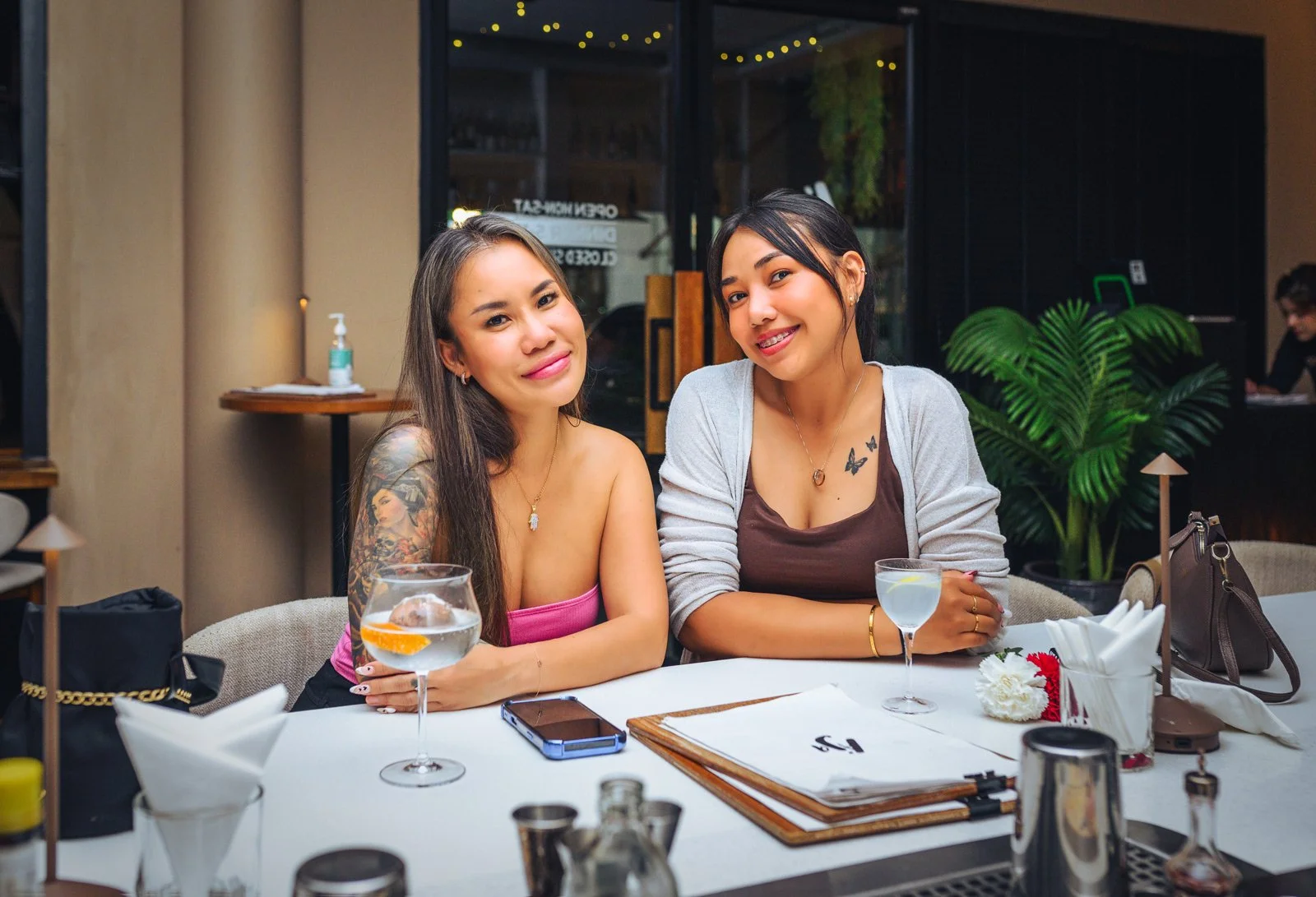 Two women sitting at a restaurant table, smiling at the camera, with drinks and a flower on the table, and a lush plant in the background.