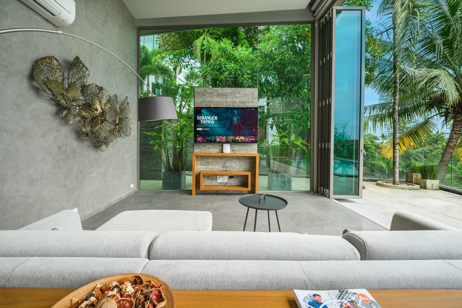 Modern living room with a large white sectional sofa, a small black side table, and a flat-screen TV showing streaming content, with a view of lush green trees outside through glass doors and windows.
