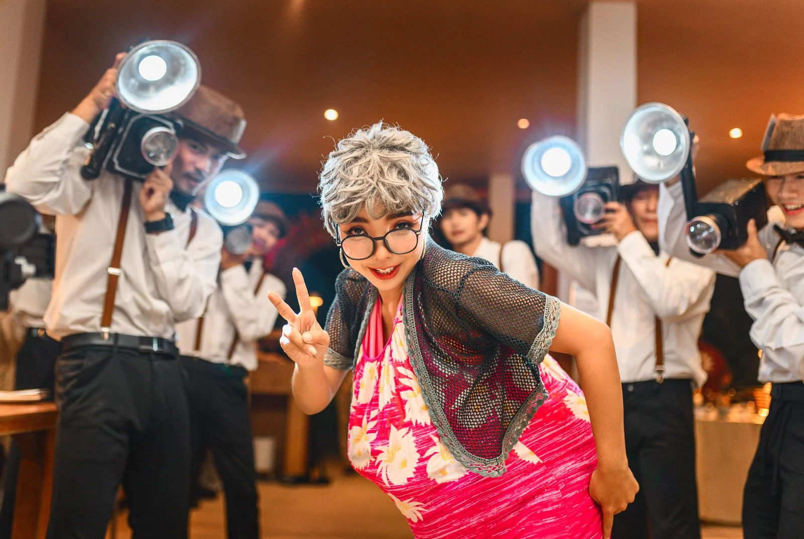 A woman with gray hair, glasses, and a bright pink floral dress is smiling and posing with a peace sign at a party. She is surrounded by people holding vintage cameras, dressed in white shirts and suspenders, taking photos of her. The scene is lively