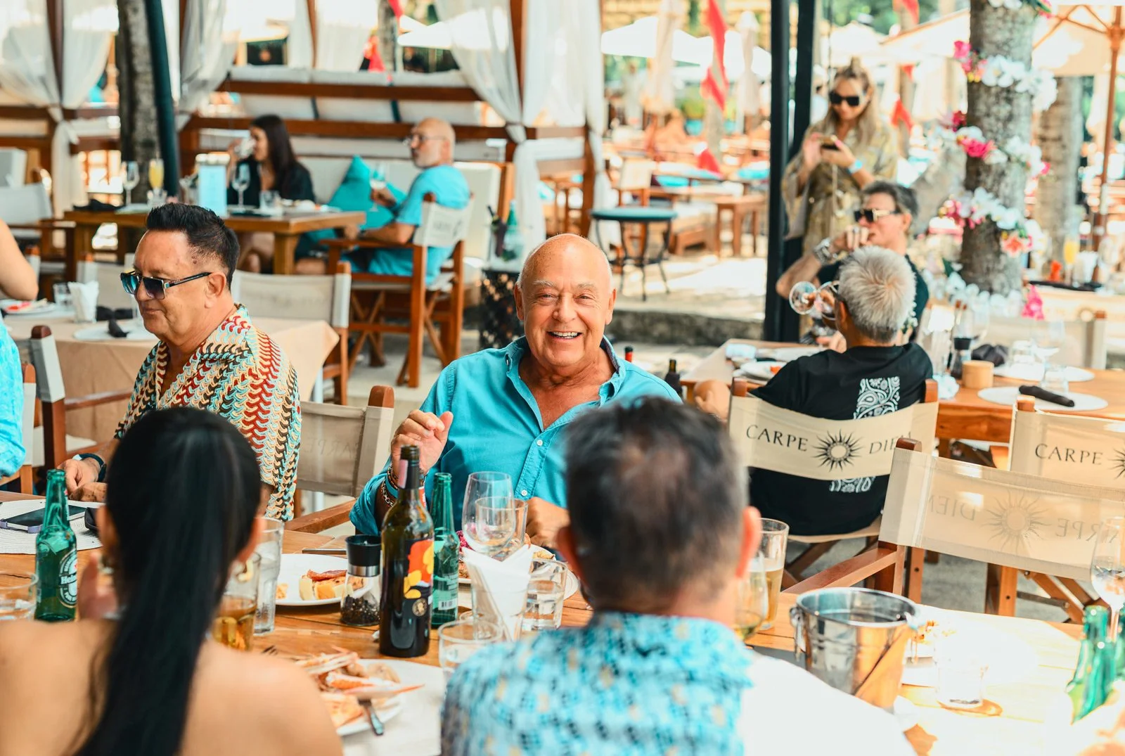 People dining at a beachside restaurant, smiling and enjoying food and drinks on a sunny day.