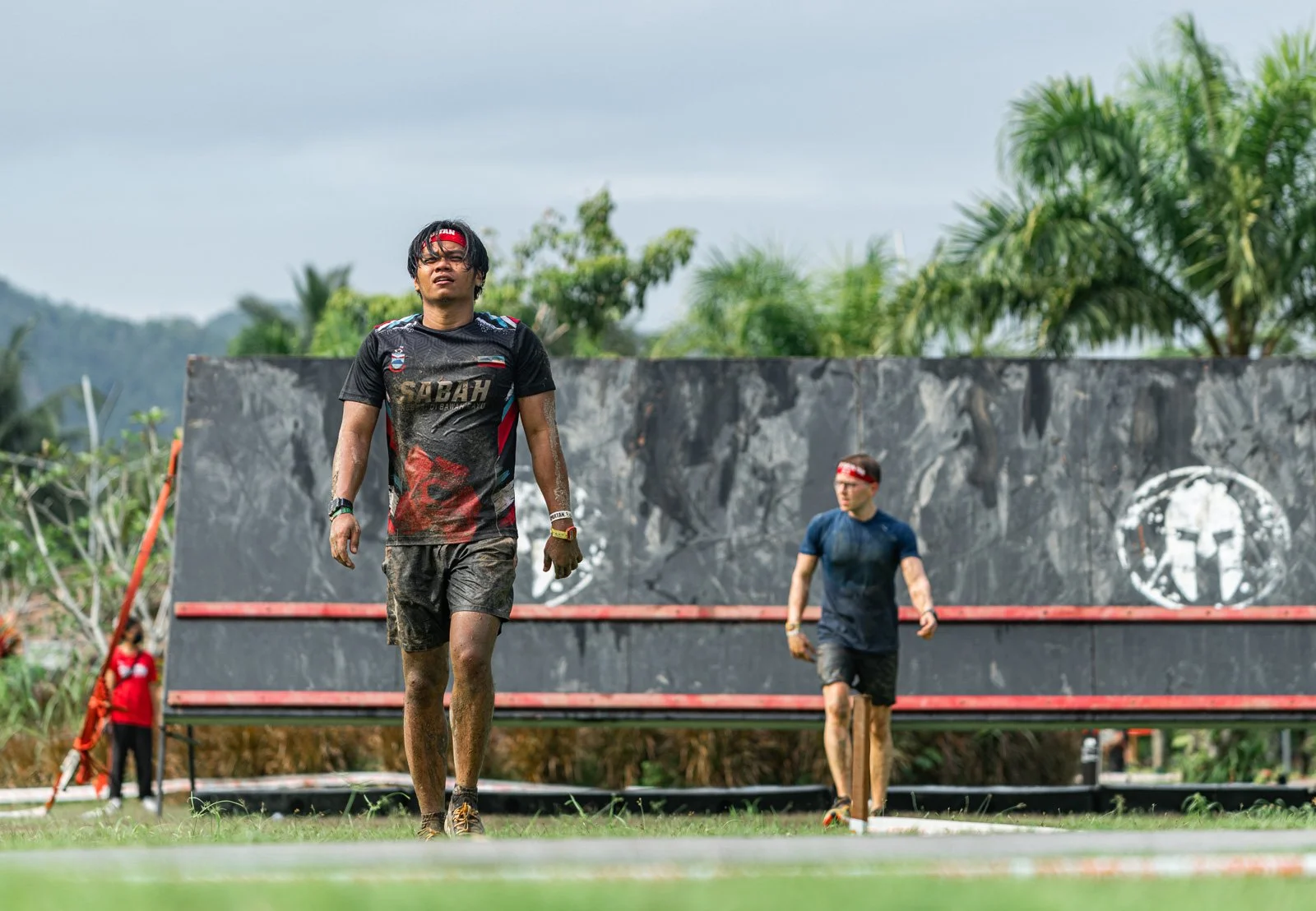 Two men walking through a muddy obstacle course outdoors, with a large black wall obstacle in the background, surrounded by tropical greenery.