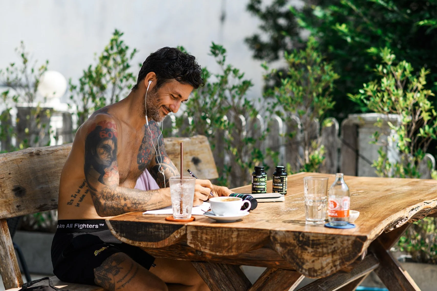 A tattooed man with dark, curly hair and a beard sitting at an outdoor wooden table, writing in a notebook with a pen. He is shirtless, wearing black shorts, and has earbuds in his ears. The table has a clear glass, a white coffee cup on a saucer, a 