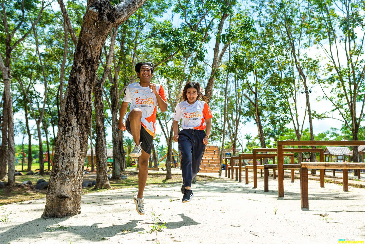 Two young people, a man and a woman, running and jumping outdoors in a park with trees, benches, and a playground in the background. They are wearing matching sports shirts and smiling.