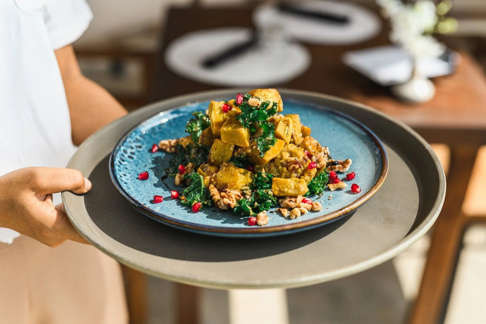 A person serving a plate of vegan curry with yellow tofu, kale, pomegranate seeds, and chopped nuts on a blue ceramic plate