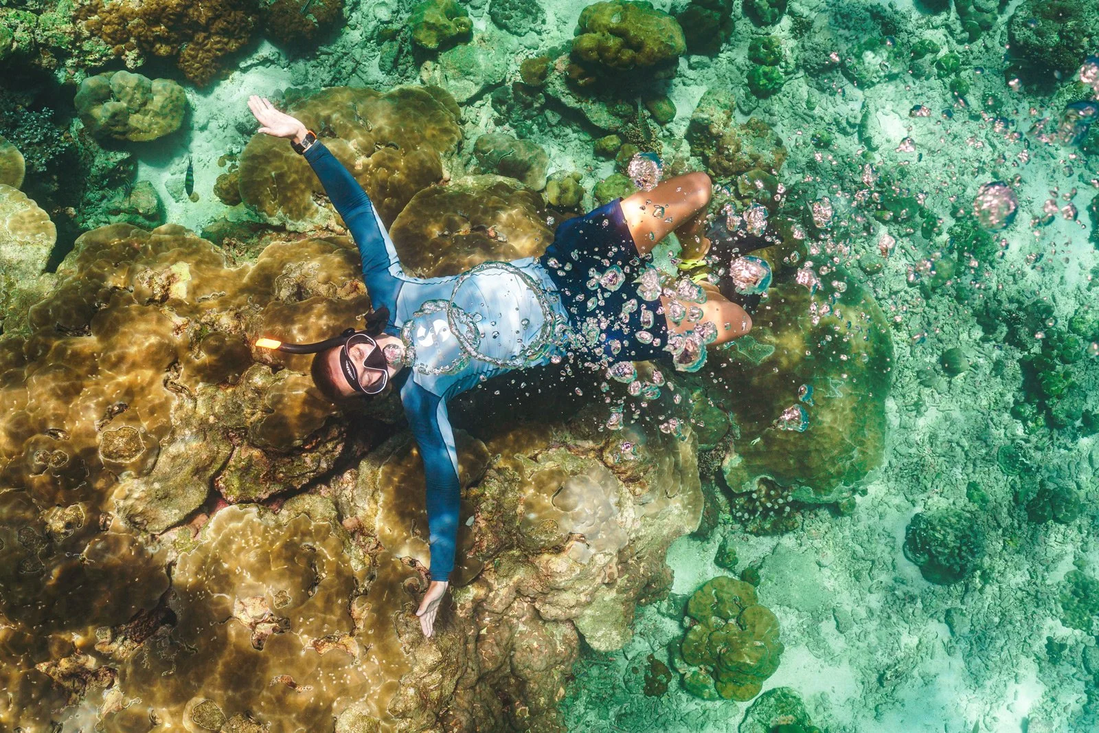 A person snorkeling underwater over a coral reef, wearing a mask, snorkel, and wetsuit, surrounded by coral and bubbles.