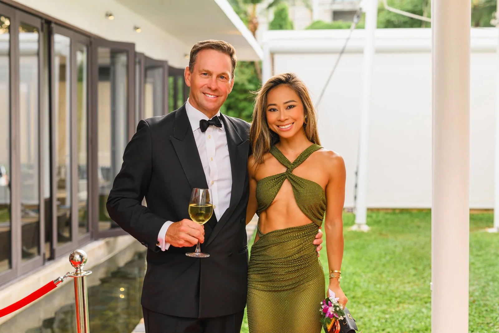 A man in a black tuxedo holding a glass of white wine stands next to a woman in a green dress holding a bouquet, both smiling at an outdoor event.