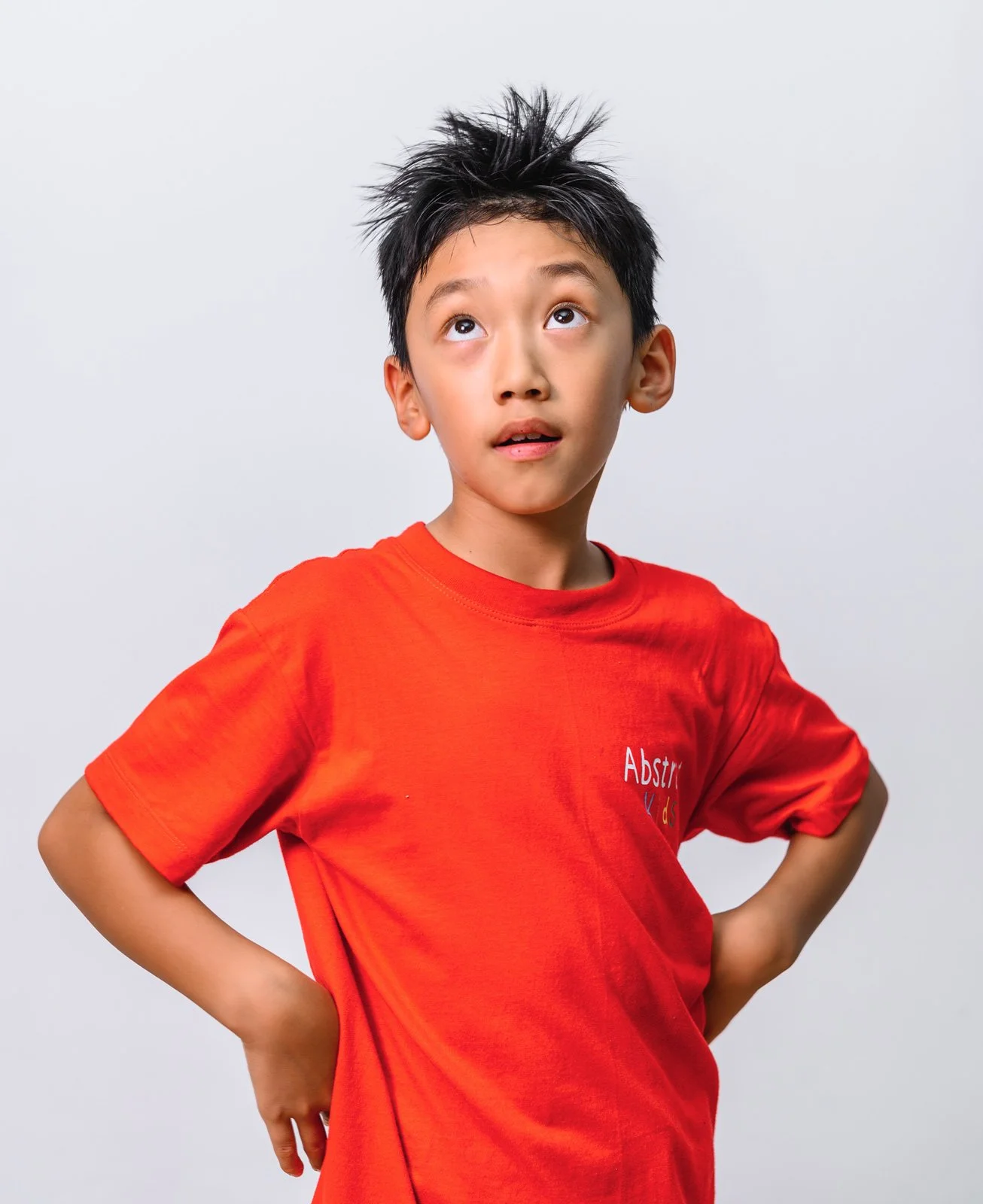 A young boy with spiky black hair looking up with a curious expression, wearing a red t-shirt with a logo that says 'Abstr' and has his hands on his hips, standing against a plain light gray background.