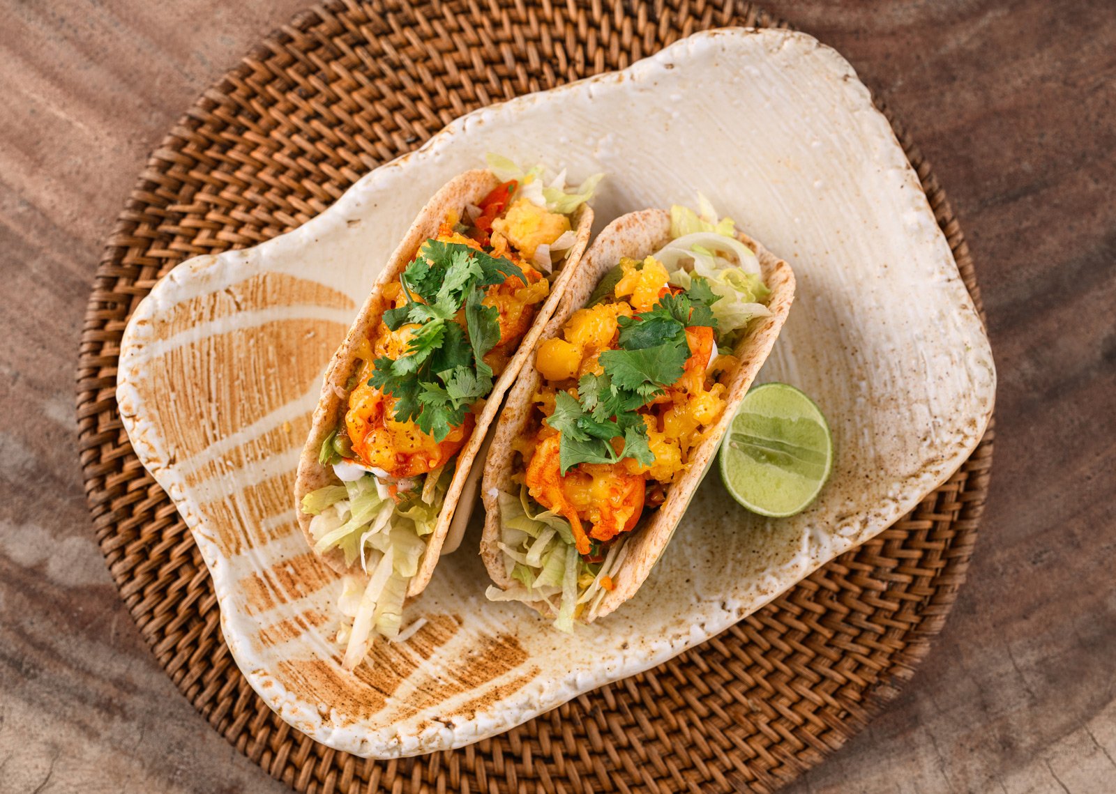 Two fish tacos with shredded lettuce, yellow sauce, cilantro, and sliced lime, served on a ceramic plate on a woven placemat.