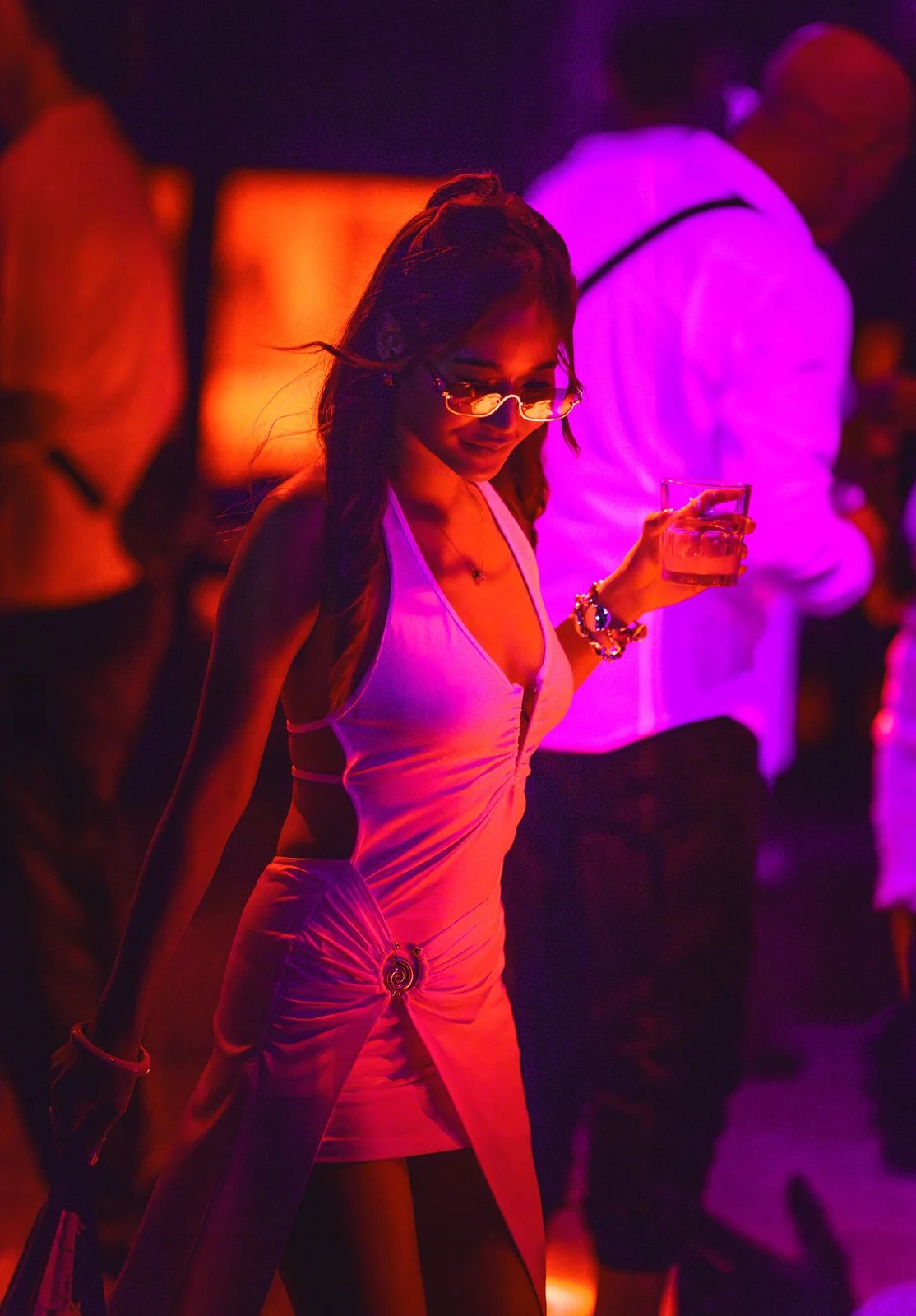 A woman at a nightclub holding a glass of drink, wearing a pink dress, with purple and orange neon lights illuminating the scene.