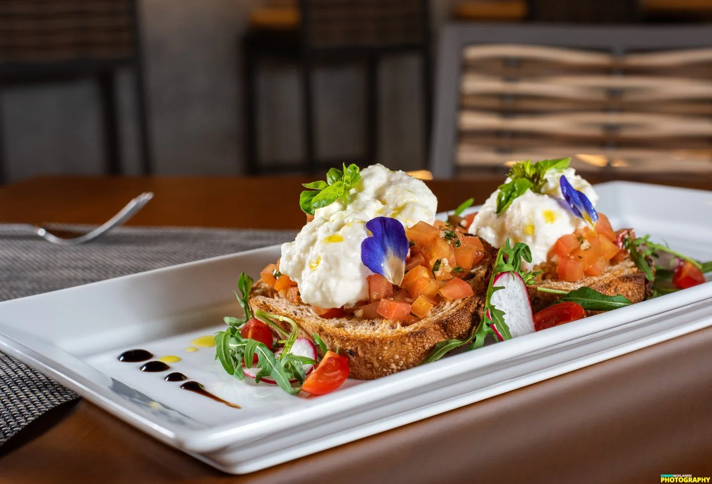 Two servings of bruschetta topped with whipped cheese, diced tomatoes, basil leaves, and edible flower petals, garnished with greens and drizzled balsamic glaze, on a white rectangular plate.