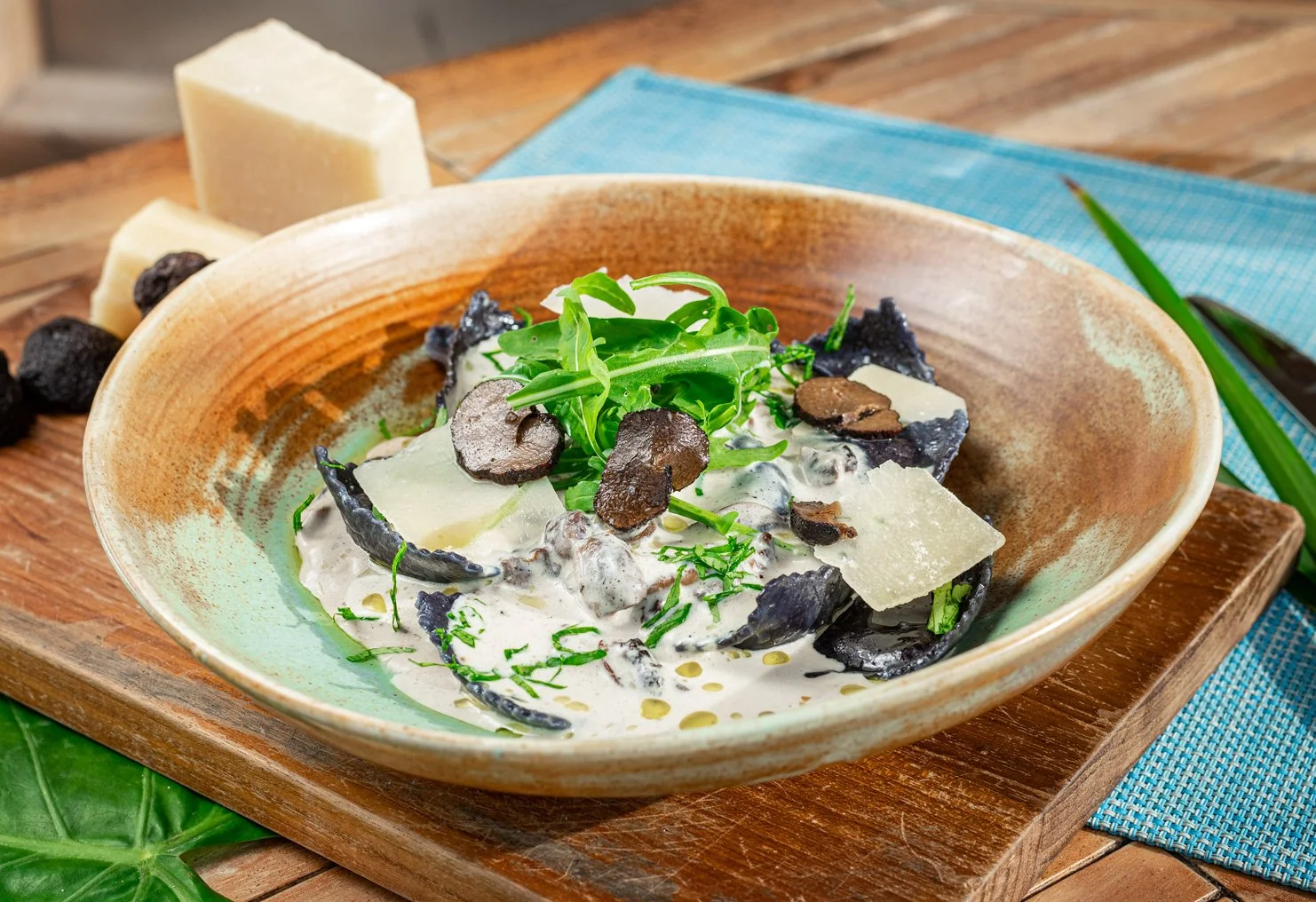 A bowl of creamy pasta with black truffle slices, topped with fresh herbs, served on a rustic wooden table with cheese and a blue napkin nearby.
