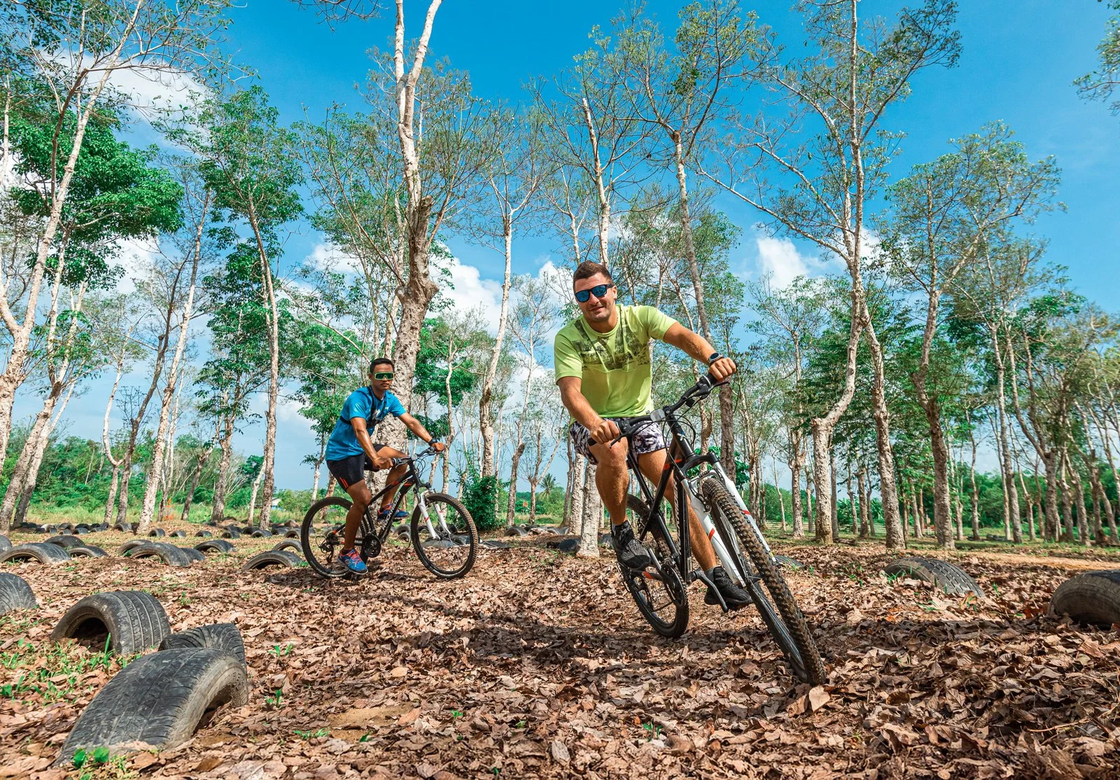 Two men riding mountain bikes on a leaf-covered trail in a forest with tall, leafless trees and a bright blue sky.