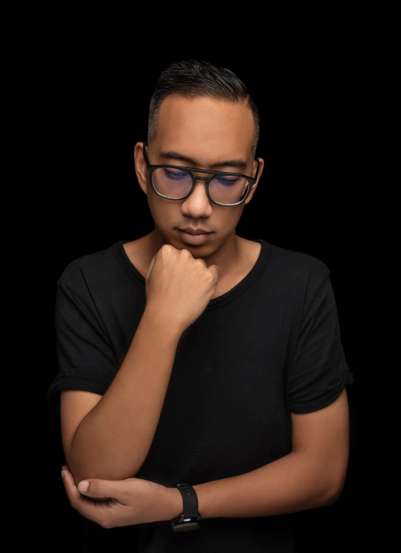 A young man with short black hair wearing glasses and a black t-shirt, standing against a black background, with his hand resting on his chin in a thoughtful pose.