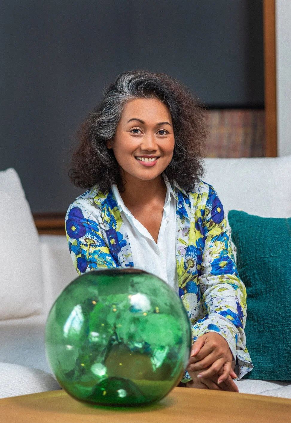 A woman with curly, salt-and-pepper hair smiling, sitting on a sofa with a green glass bowl on the table in front of her.