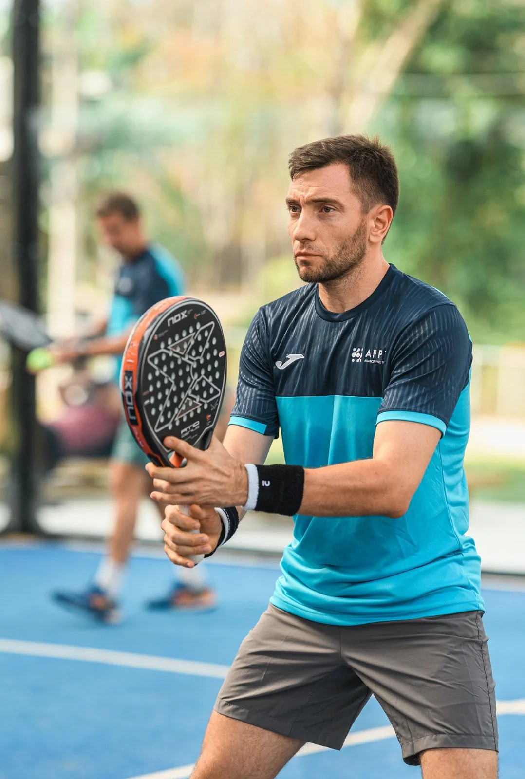 A man in a blue sports shirt playing padel tennis, holding a black and orange paddle on an outdoor court with trees in the background.