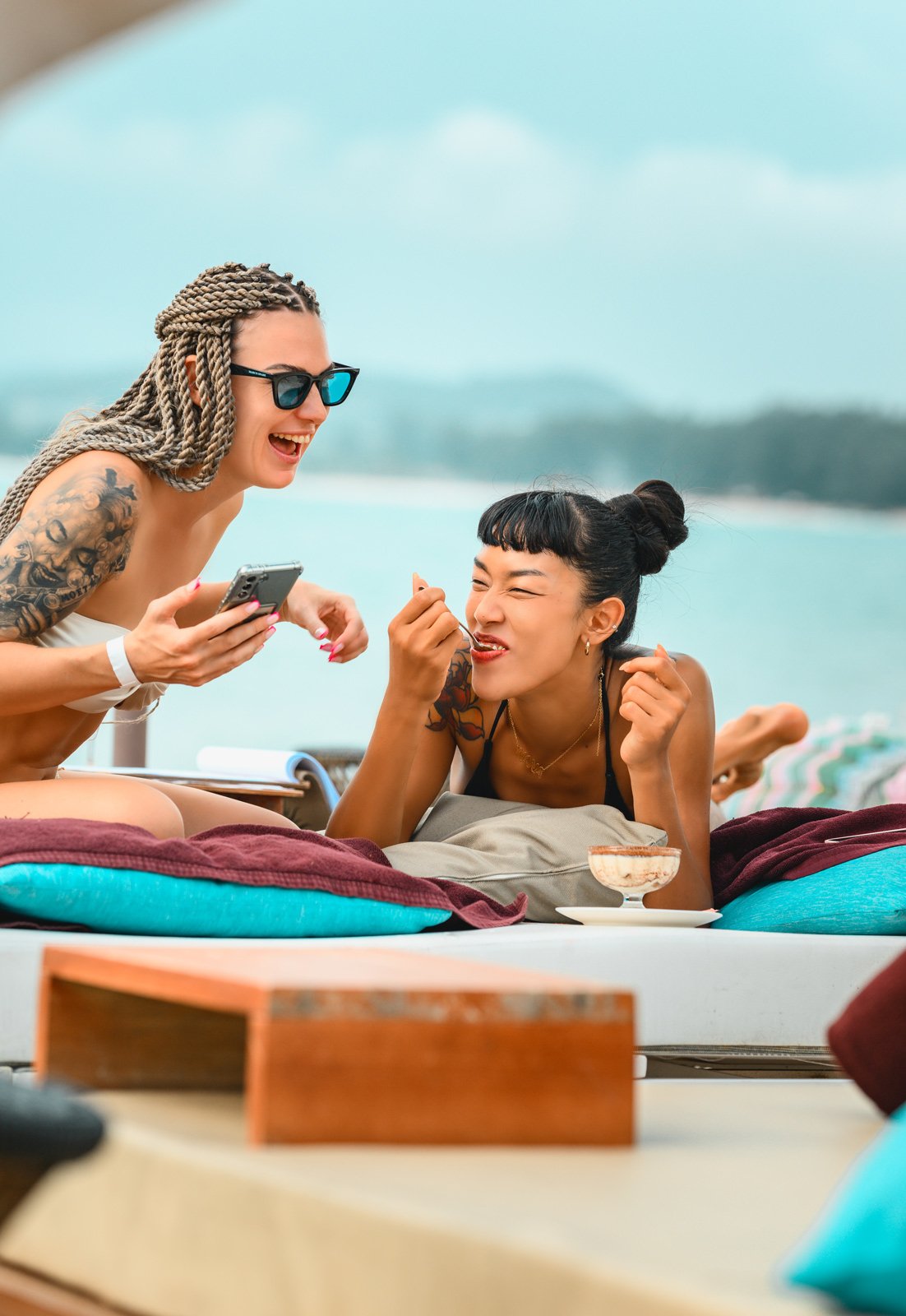 Two women relaxing at the beach, one showing something on her phone to the other, with a drink in a glass nearby, beach and water in the background.