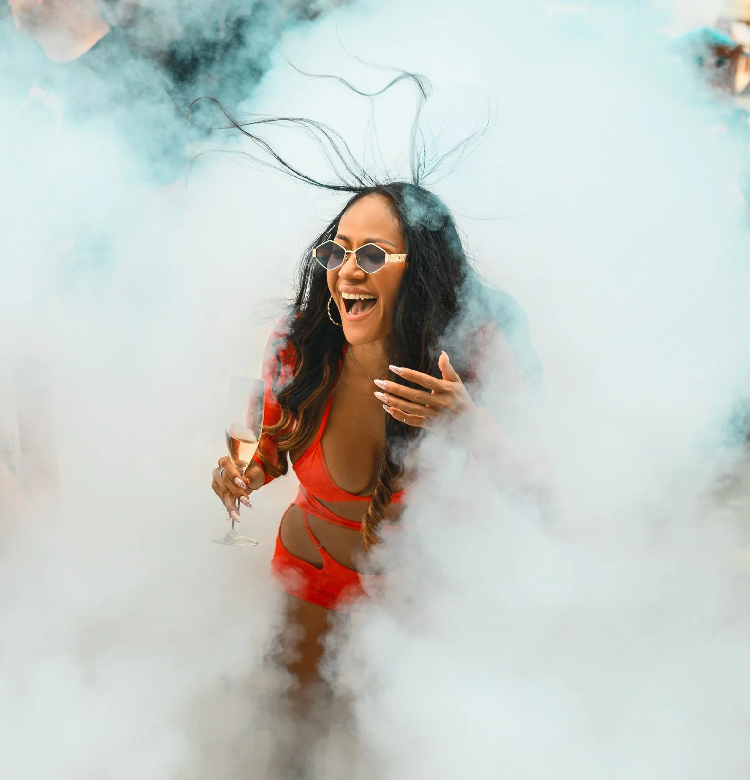 Woman in red swimsuit wearing sunglasses, surrounded by smoke or fog, holding a champagne glass, smiling and laughing.