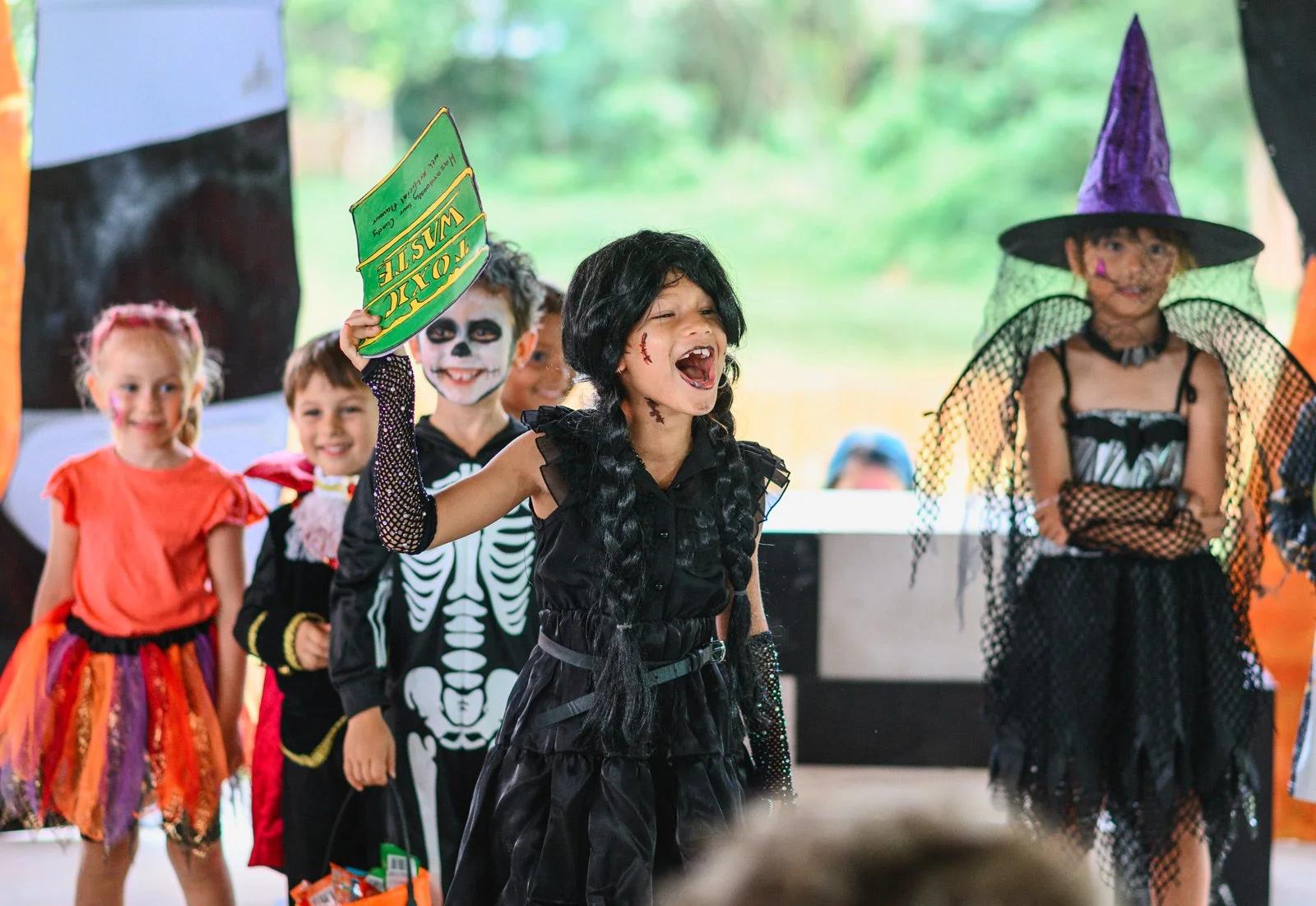 Children dressed in Halloween costumes, including a girl in a black dress with fake scars, a skeleton costume, a witch costume, and a princess dress, standing together at a Halloween celebration.