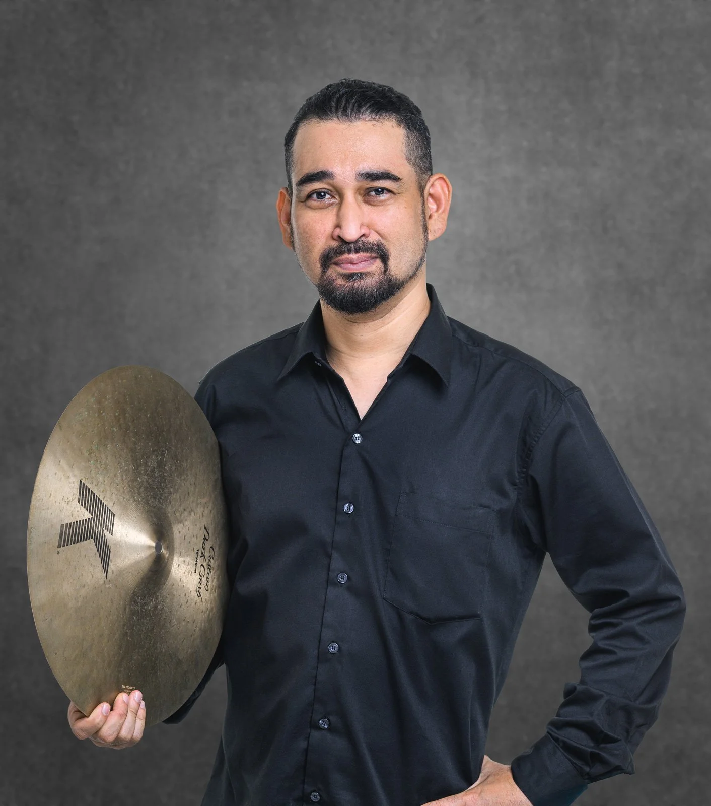 A man with dark hair and a goatee holding a cymbal, standing against a gray background. He is wearing a black button-up shirt and has a confident expression.