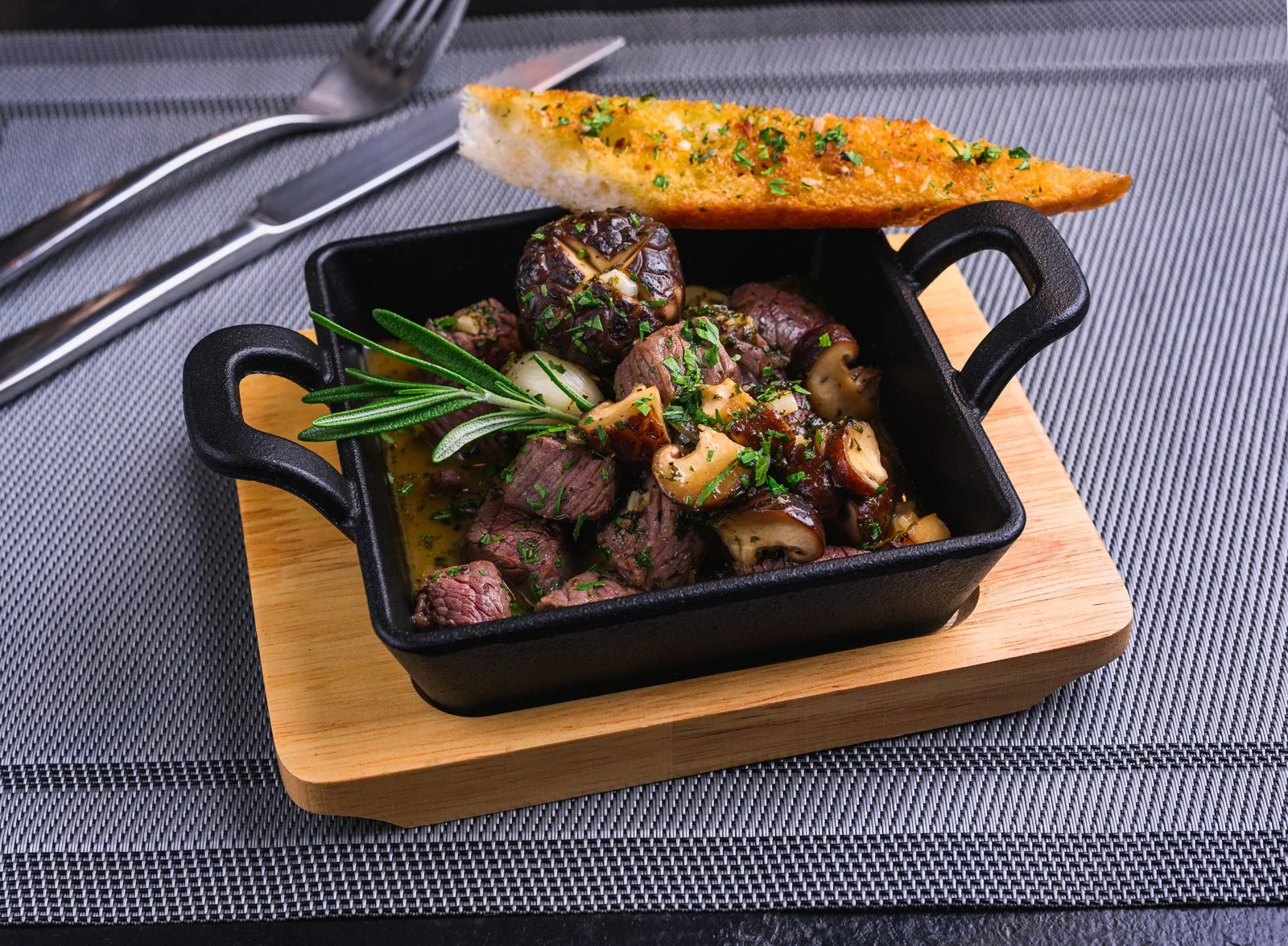 A small black cast iron skillet on a wooden board, filled with cooked beef and mushroom stew garnished with chopped herbs and a sprig of rosemary, with a piece of toasted bread on the edge of the skillet. The skillet is on a textured gray tablecloth 