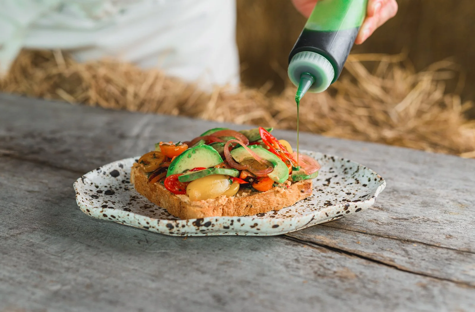 A slice of bread topped with sliced vegetables like tomatoes, cucumbers, and olives, is being drizzled with green sauce from a squeeze bottle.