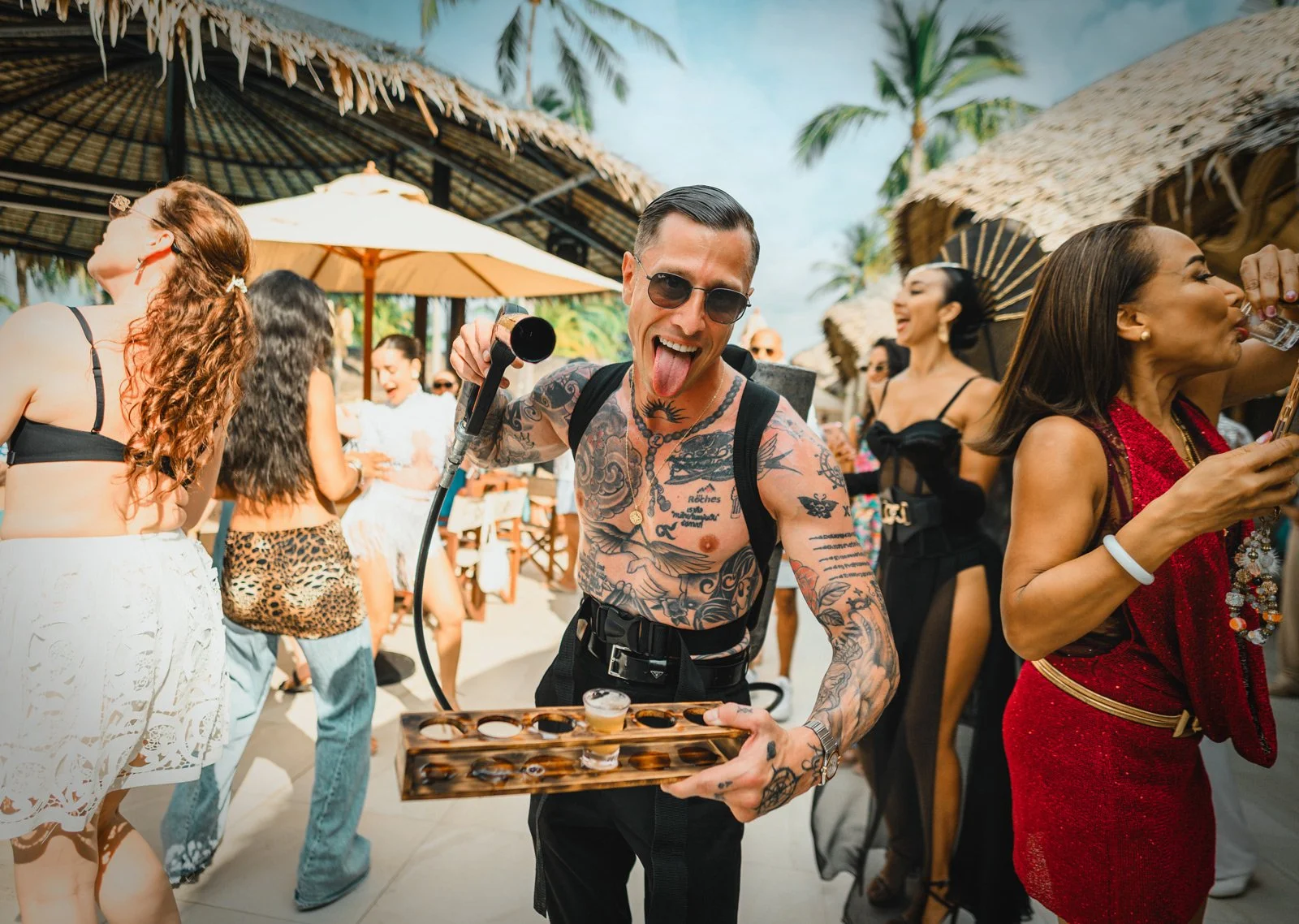 A man with tattoos and sunglasses dancing and holding a shot glass at a lively outdoor party with people in summer attire, tiki umbrellas, and palm trees.